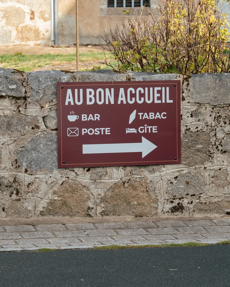 Le panneau d'Au Bon Accueil sur un muret : bar, tabac, poste et gîte, avec une flèche